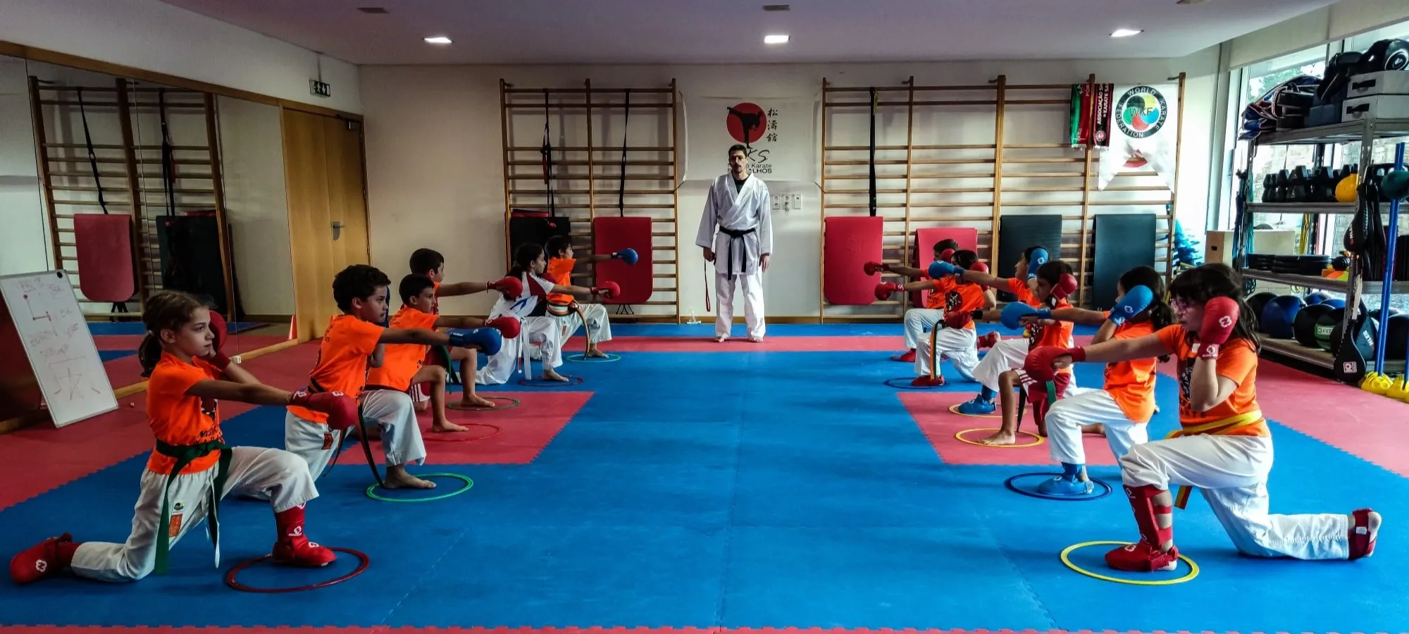 Treinador observa crianças de t-shirt laranja a treinar socos com luvas no tatami
